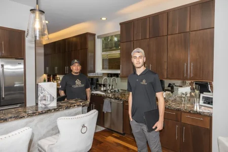 Two men stand in a modern kitchen with dark wood cabinets, marble countertops, and stainless steel appliances. One man stands behind the counter, while the other stands in front, holding a black folder.