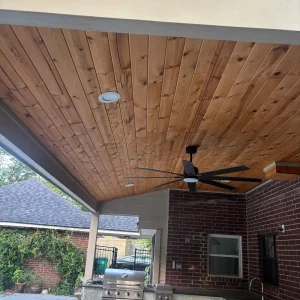 A covered outdoor patio with a wooden ceiling, ceiling fan, built-in grill, and sink. A brick wall and a pool are visible in the background, with trees and a nearby house outside the patio area.