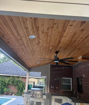 A covered outdoor patio with a wooden ceiling, ceiling fan, built-in grill, and sink. A brick wall and a pool are visible in the background, with trees and a nearby house outside the patio area.