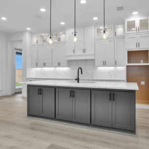 Modern kitchen with a large island featuring dark gray cabinets, white countertops, pendant lights, white upper cabinets, marble backsplash, light wood flooring, and large windows letting in natural light.